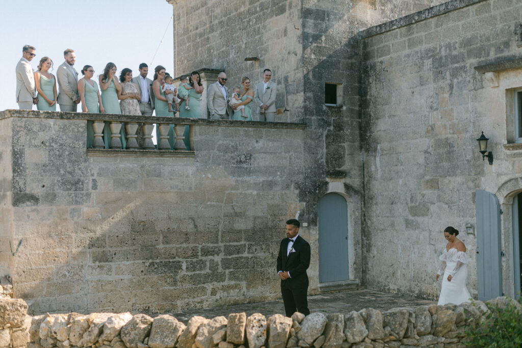 A Puglian wedding affair unfolds as the groom stands with bated breath, about to turn for the first look at his radiant bride amidst the olive groves
