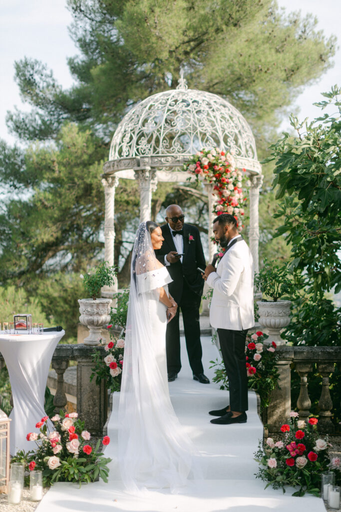 Couple exchanging vows under a floral arch at Château Saint-Georges, with guests and the Riviera's landscape in the background, epitomizing their French-Riviera-St-Georges-Wedding