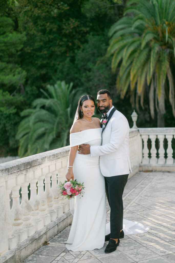 Newlyweds embracing in the gardens of Château Saint-Georges, the Mediterranean Sea glittering in the distance, a serene moment from their French-Riviera-St-Georges-Wedding